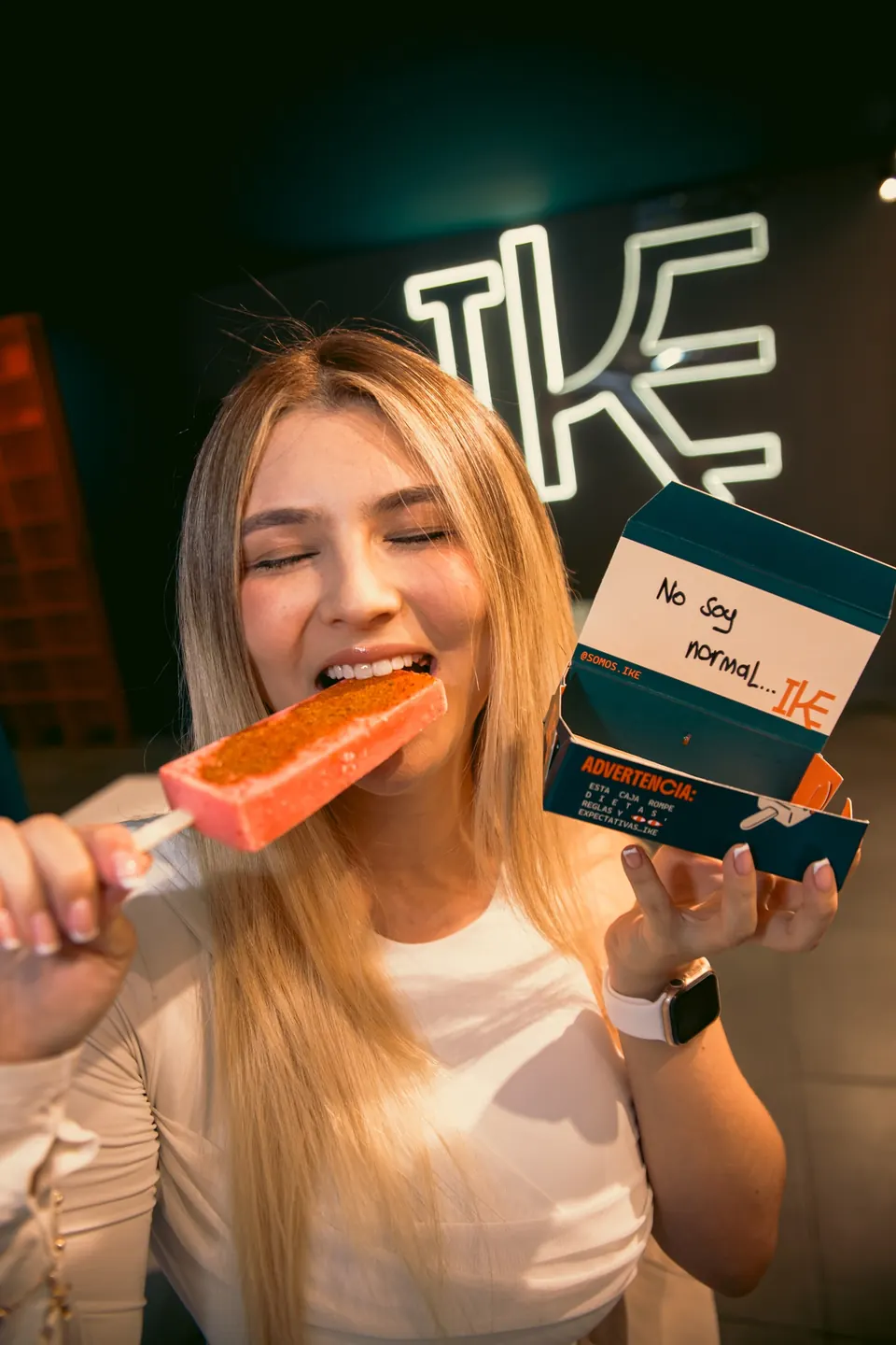 Joven mordiendo una paleta frutal IKE y mostrando la caja "No soy normal" frente al neón de IKE en Medellín